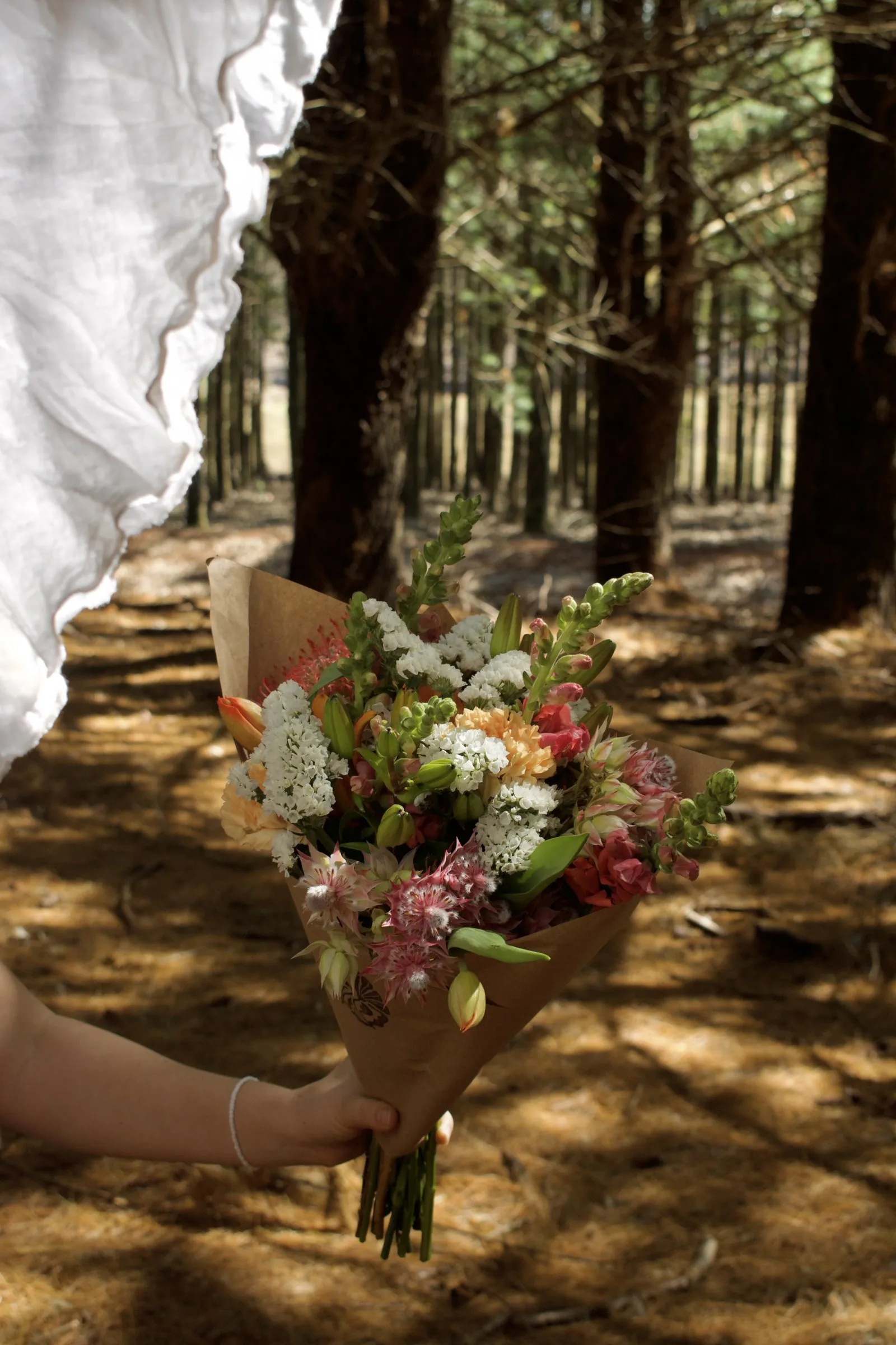 A hand holding a kraft-wrapped bouquet of proteas, snapdragons and lilies in a pine forest