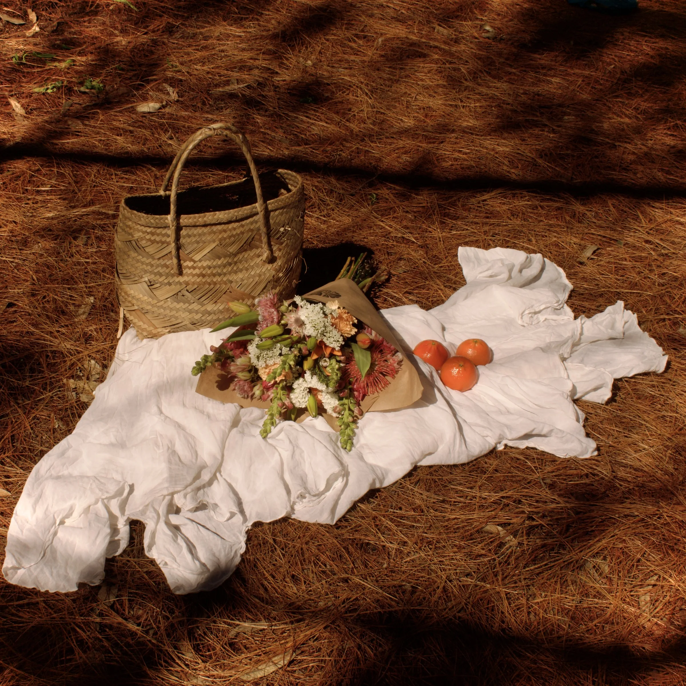A wrapped bouquet, woven basket and three oranges laid on white linen among pine needles