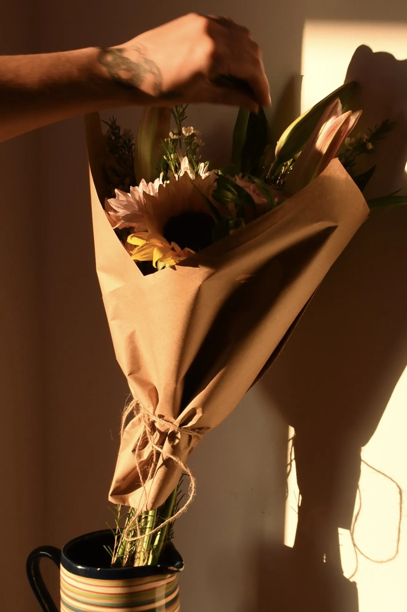 A sunflower bouquet in kraft paper being lifted from a striped mug in evening light