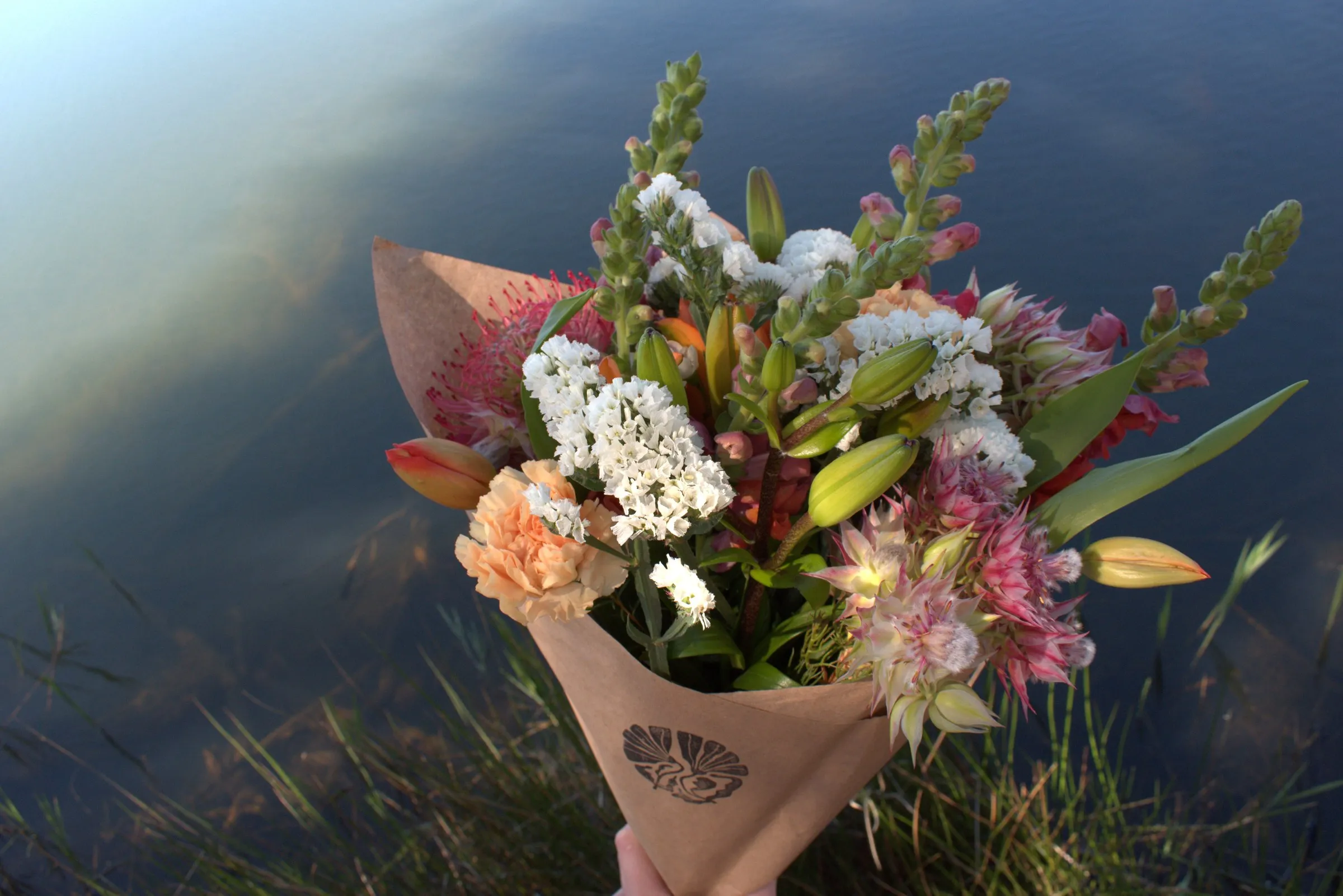 A kraft-wrapped bouquet held above still water at golden hour, Eden stamp on the paper